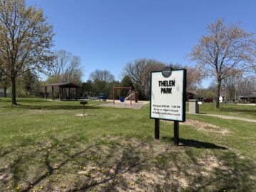 Picture of a sign for Thelen Park with red and blue playground equipment, an open air pavilion and a grill in the background.