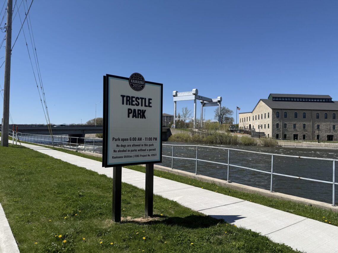 Picture of a sign for Trestle Park, with the Veterans Memorial Bridge and Ahlstrom-Thilmany building in the background