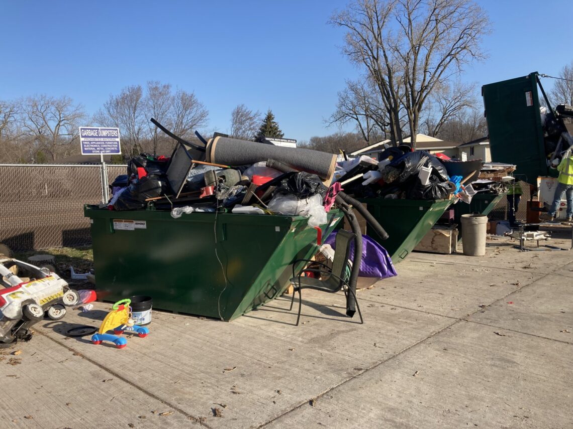 picture of two green dumpsters overflowing with carpet, wood, furniture, toys and other various items. Items are stacked alongside the dumpsters as well.