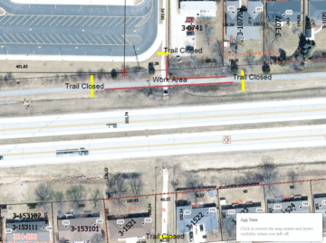 Aerial view of a road with three "Trail Closed" signs and one "Work Area" sign positioned across pathways adjacent to the road. This indicates temporary closures due to construction or maintenance work. Residential buildings and parking areas are nearby.